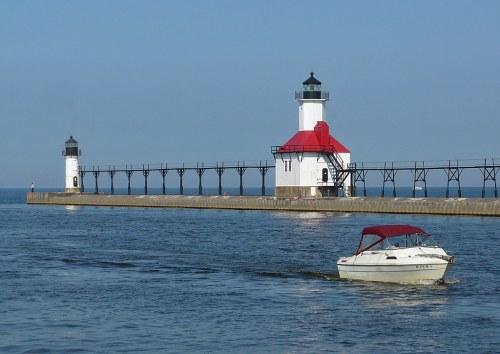 A boat sails past two lighthouses on a pier, with calm blue waters and a clear sky in the background.