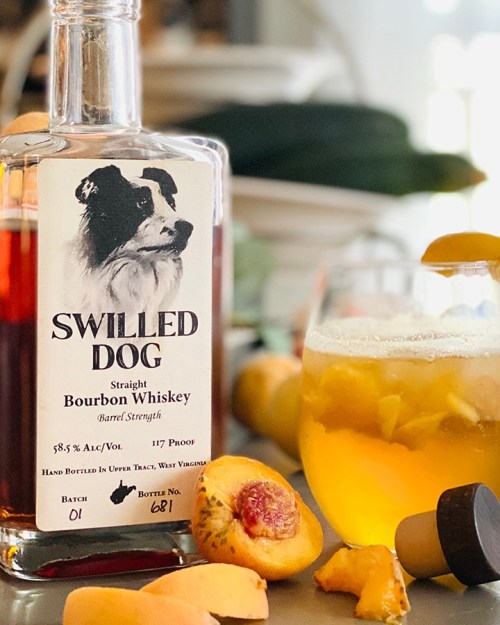 A bottle of Swilled Dog bourbon whiskey next to a glass of whiskey and fresh peaches on a table.