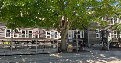 A rustic restaurant with outdoor seating under a large tree, featuring a wooden facade and American flags.