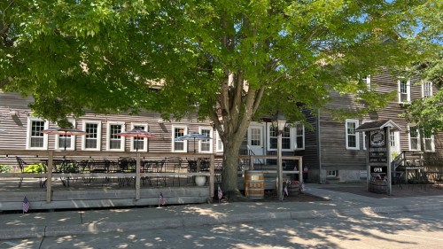 A rustic wooden building with outdoor seating under a large tree, featuring umbrellas and a sign that says "Open."