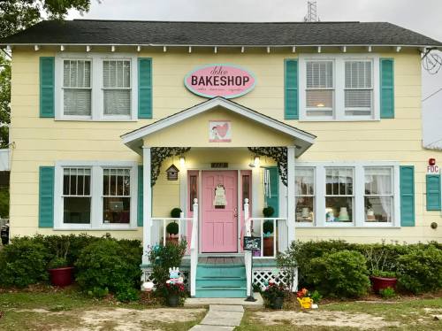 A charming yellow bakeshop with teal shutters and a pink door, featuring a welcoming porch and decorative plants.