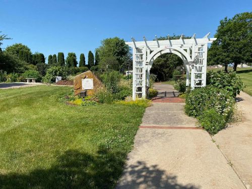 A white arbor frames a pathway in a lush garden, with greenery and a sign visible on the left. Clear blue sky above.