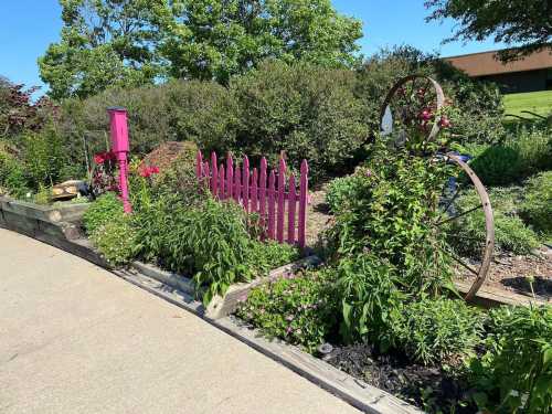 A vibrant garden featuring a pink fence, flowers, and a decorative wheel against a clear blue sky.