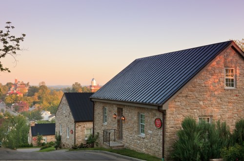 A stone house with a metal roof on a hillside, overlooking a town at sunrise with soft pastel skies.