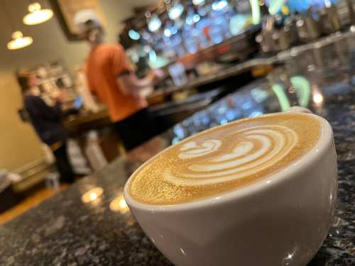 A close-up of a latte with intricate foam art, with baristas working in the background of a cozy café.