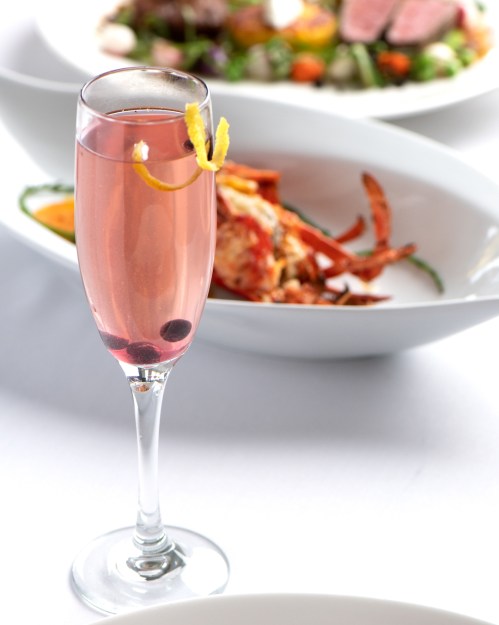 A pink cocktail garnished with a lemon twist and a berry, with a plate of seafood in the background.