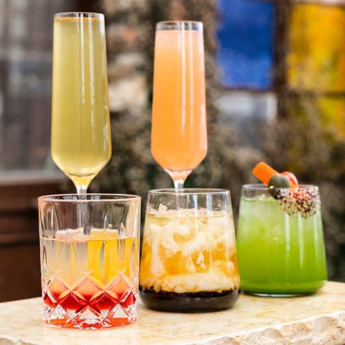 Four colorful cocktails in elegant glasses, featuring vibrant hues and garnishes, displayed on a stone surface.