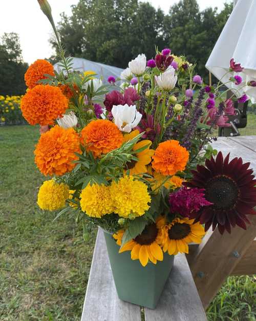 A vibrant bouquet of orange marigolds, sunflowers, and various colorful flowers in a green vase, set outdoors.