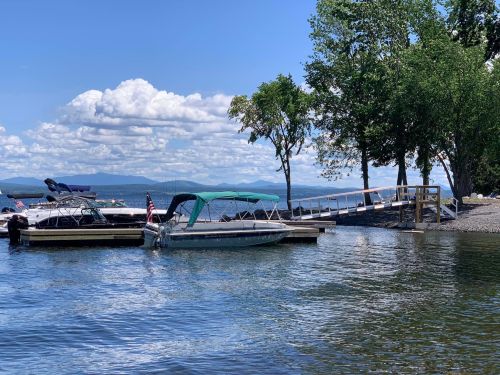 Boats docked at a lakeside with trees and a wooden ramp under a blue sky with fluffy clouds.