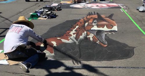 An artist creates a colorful dinosaur chalk drawing on pavement, surrounded by art supplies and other artworks.