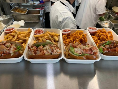 Four trays of lobster rolls with fries and curly fries, served with dipping sauces in a busy kitchen setting.