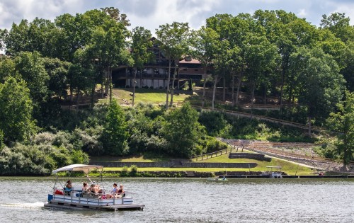 A boat with people floats on a lake, with a wooded hillside and a house in the background under a partly cloudy sky.