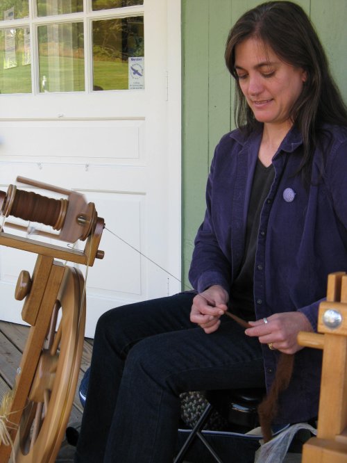 A woman sits at a spinning wheel, carefully working with fiber, focused and engaged in her craft.