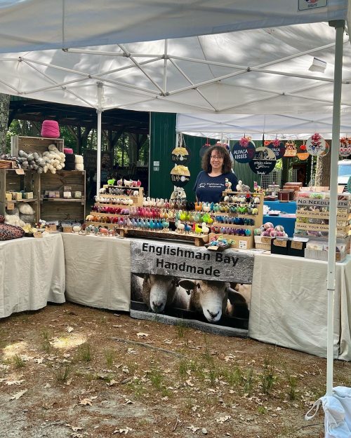 A vendor stands behind a table filled with handmade crafts at a market, with a banner reading "Englishman Bay Handmade."
