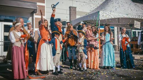A group of people in historical costumes celebrate with confetti and a ribbon-cutting ceremony outside a building.