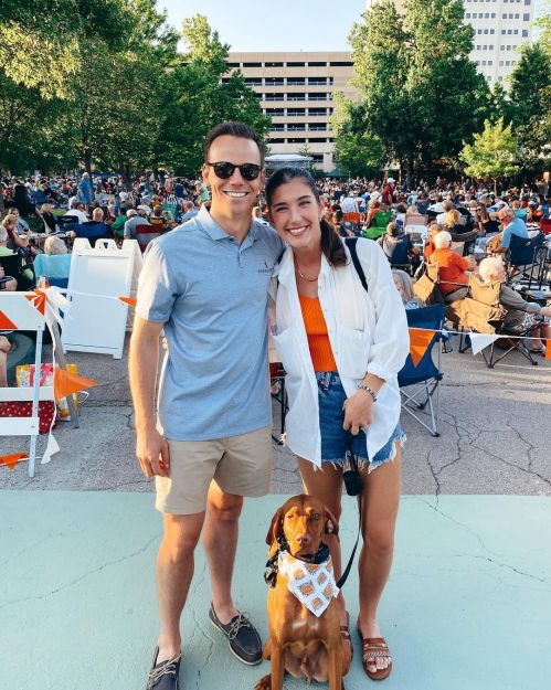 A young couple poses with a dog at an outdoor event, surrounded by a crowd and trees in the background.