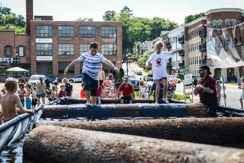 Children balance on logs in a water-filled area during a summer festival, with a crowd enjoying the event nearby.