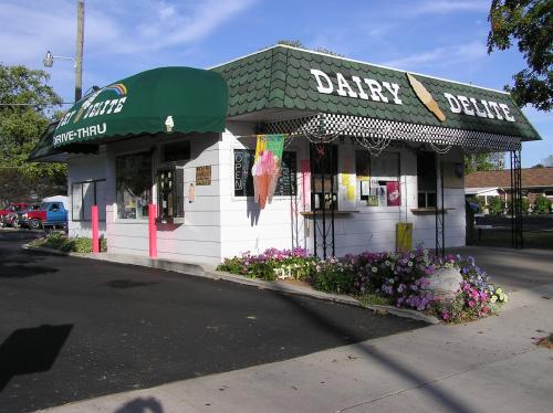 A small ice cream shop with a green awning, colorful flowers, and a drive-thru sign on a sunny day.