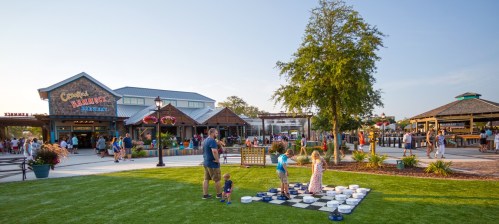 A family enjoys a sunny day at a marketplace, with children playing checkers on a large board in the foreground.
