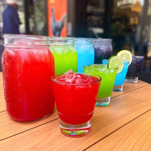 A colorful array of drinks in various glasses, including red, green, blue, and purple, on a wooden table.