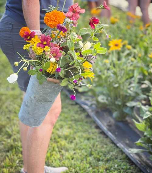 A person holds a metal bucket filled with vibrant, colorful flowers in a sunny garden setting.