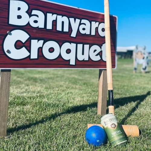 A sign for "Barnyard Croquet" with croquet mallets and balls on green grass, featuring a farm backdrop.