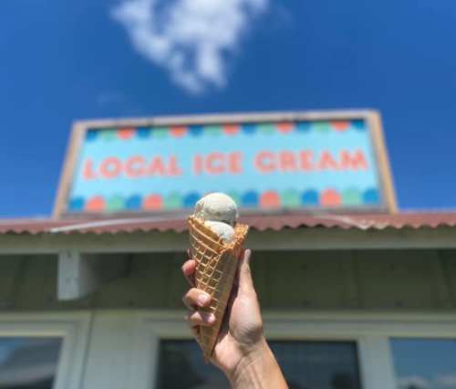 A hand holds a cone with three scoops of ice cream in front of a colorful "Local Ice Cream" sign against a blue sky.