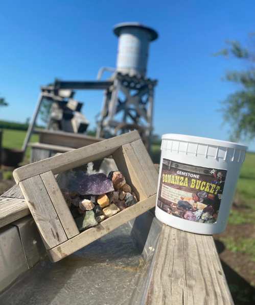 A bucket labeled "Gemstone Bonanza" next to a wooden frame displaying colorful gemstones, with a water feature in the background.