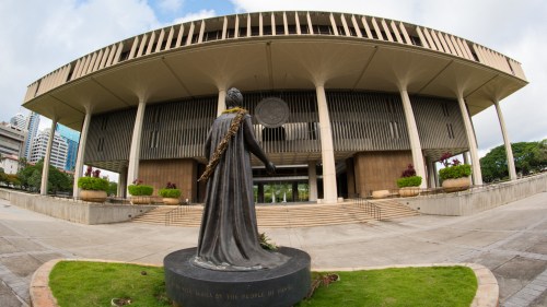 Statue of a figure in front of a modern government building with a landscaped area and cloudy sky.
