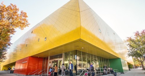 A modern building with a striking yellow and green facade, surrounded by trees and people at the entrance.