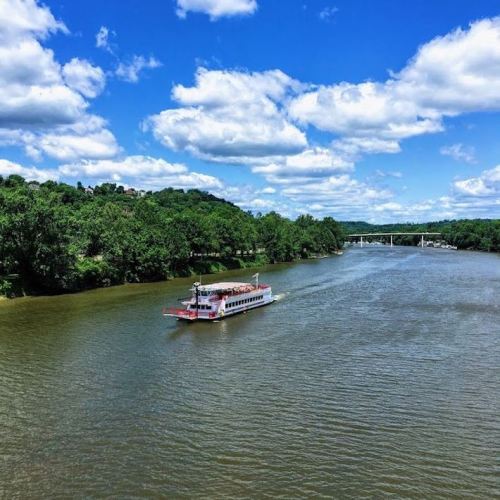 A boat glides along a river surrounded by lush greenery and a bright blue sky with fluffy clouds.