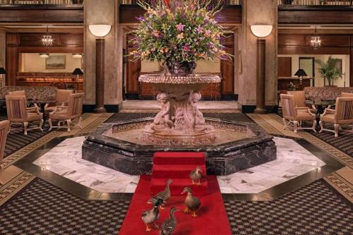 A grand hotel lobby featuring a floral fountain, elegant seating, and a red carpet with four ducks walking across it.