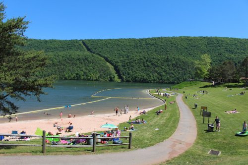 A sunny beach scene with people relaxing on the sand and grass, surrounded by green hills and a calm lake.