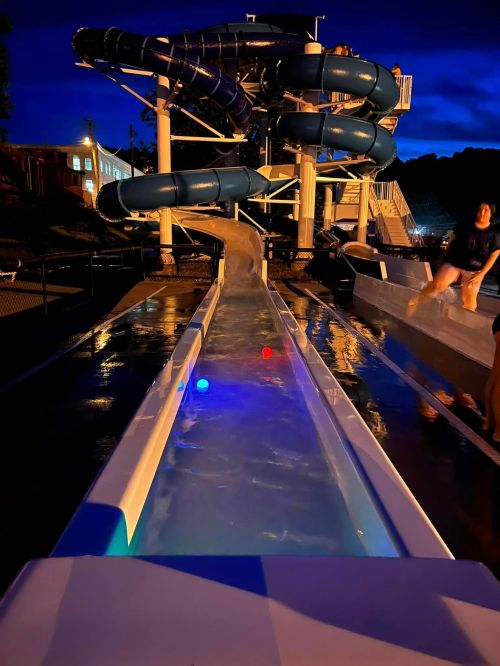 A water slide at night, illuminated by blue lights, with a clear water path and people enjoying the ride.