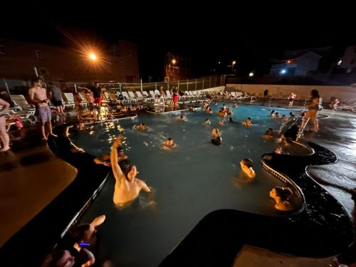 A lively night scene at a pool with people swimming and relaxing around the water, illuminated by lights.