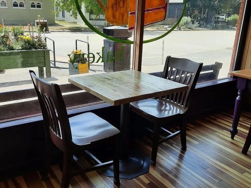 A small wooden table with two chairs by a window, overlooking a garden and street outside.
