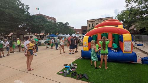 A lively outdoor event with families enjoying a bounce house and festive activities, surrounded by green decorations.