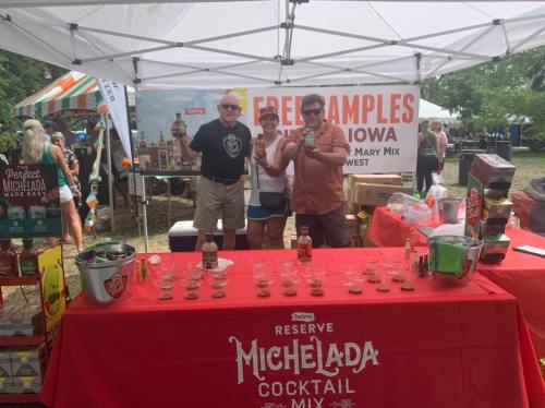 Three people pose with drinks at a booth offering free samples of Michelada cocktail mix at an outdoor event.