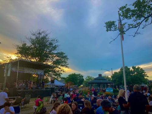 A lively outdoor event at sunset, with a stage, audience, and trees under a colorful sky.