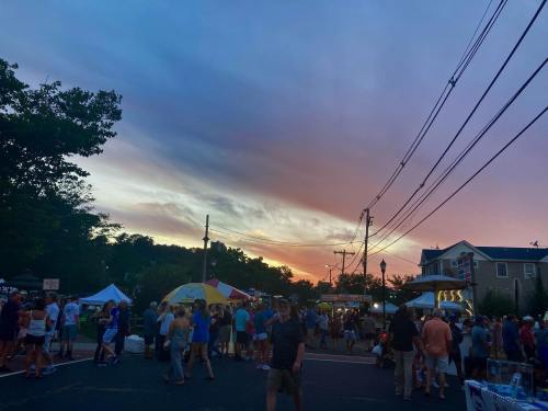 A vibrant sunset over a bustling street fair, with crowds enjoying food and festivities under colorful skies.