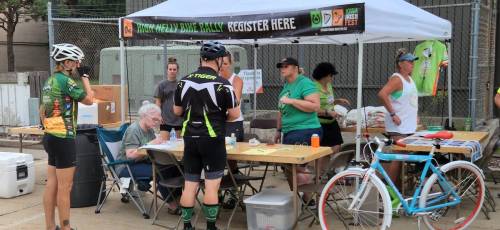 A group of people at a registration booth for a bike rally, with tables, chairs, and a banner overhead.