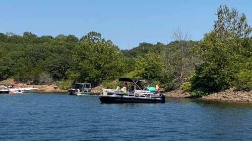 A pontoon boat with people is anchored on a calm lake surrounded by green trees and other boats in the background.
