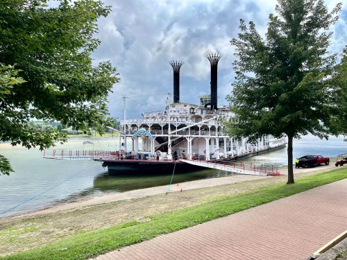 A large steamboat docked by a river, with two tall smokestacks and a ramp leading to the shore, surrounded by trees.