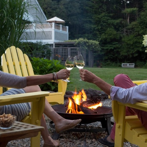 A couple toasts with glasses of wine by a fire pit, seated in yellow chairs in a lush garden setting.
