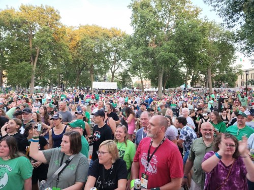 A large crowd of people in green shirts enjoying an outdoor event, with trees and tents in the background.