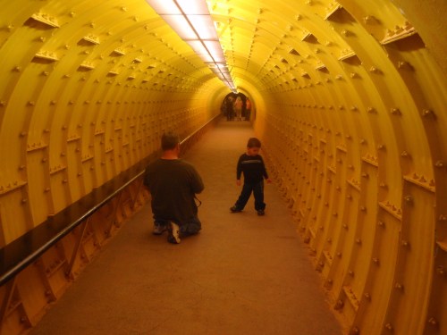 A child stands in a yellow tunnel while an adult kneels nearby, with people visible in the background.