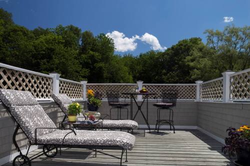 A sunny rooftop patio with lounge chairs, a small table, and potted plants, surrounded by greenery and blue skies.