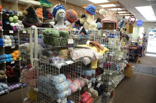 A cozy yarn shop filled with colorful yarns, knitted items, and displays on shelves and mannequins.