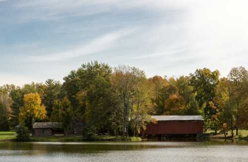 A serene landscape featuring a red covered bridge and rustic buildings surrounded by colorful autumn trees by a calm lake.