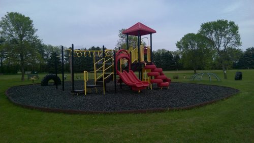 Colorful playground with slides and climbing structures, surrounded by grass and trees under a cloudy sky.